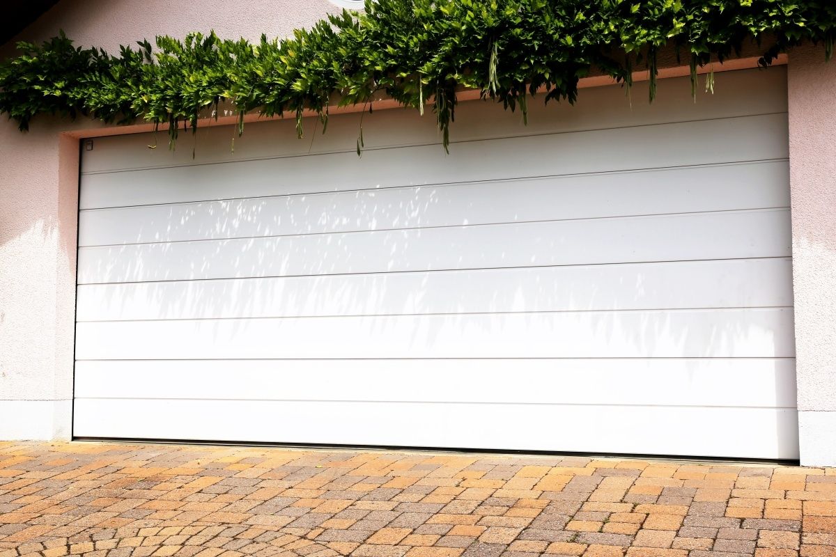 Eine Einfahrt mit einem weißen Garagentor und Grünpflanzen dadrüber